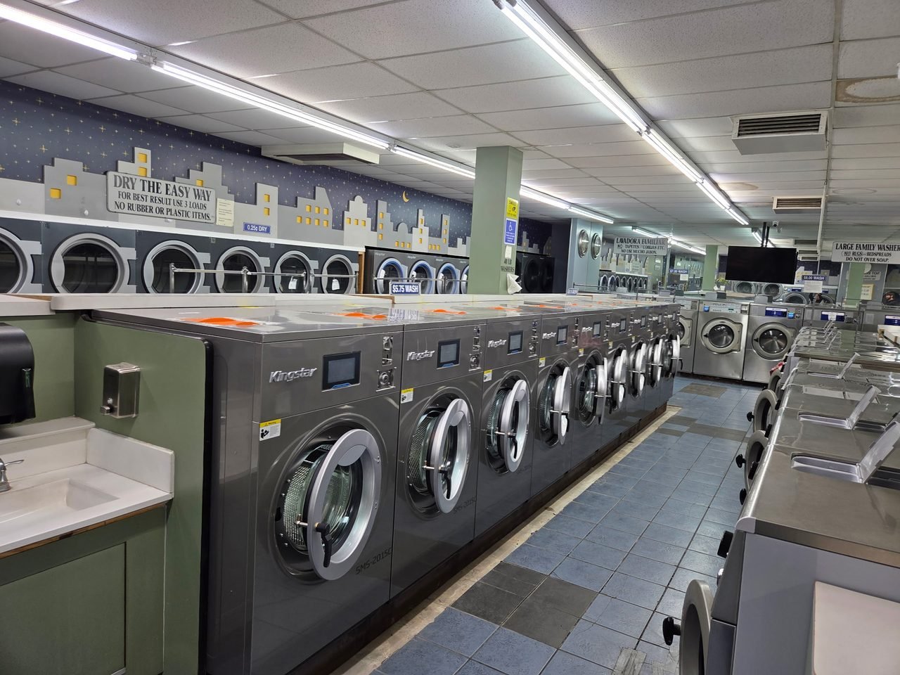 Wide view of a laundromat refitted with Kingstar SMS-2015C washers along the centre island, with existing dryer walls and folding tables kept in service around them.