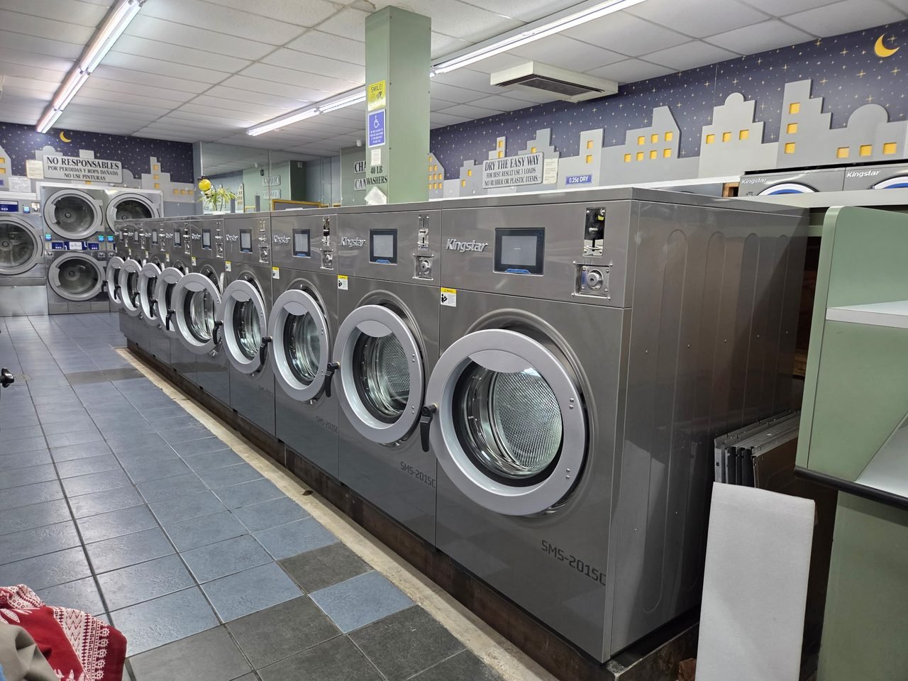 A row of installed Kingstar SMS-2015C coin-operated washers on a working laundromat floor, each with its own vend screen and coin drop under a painted city-skyline mural.